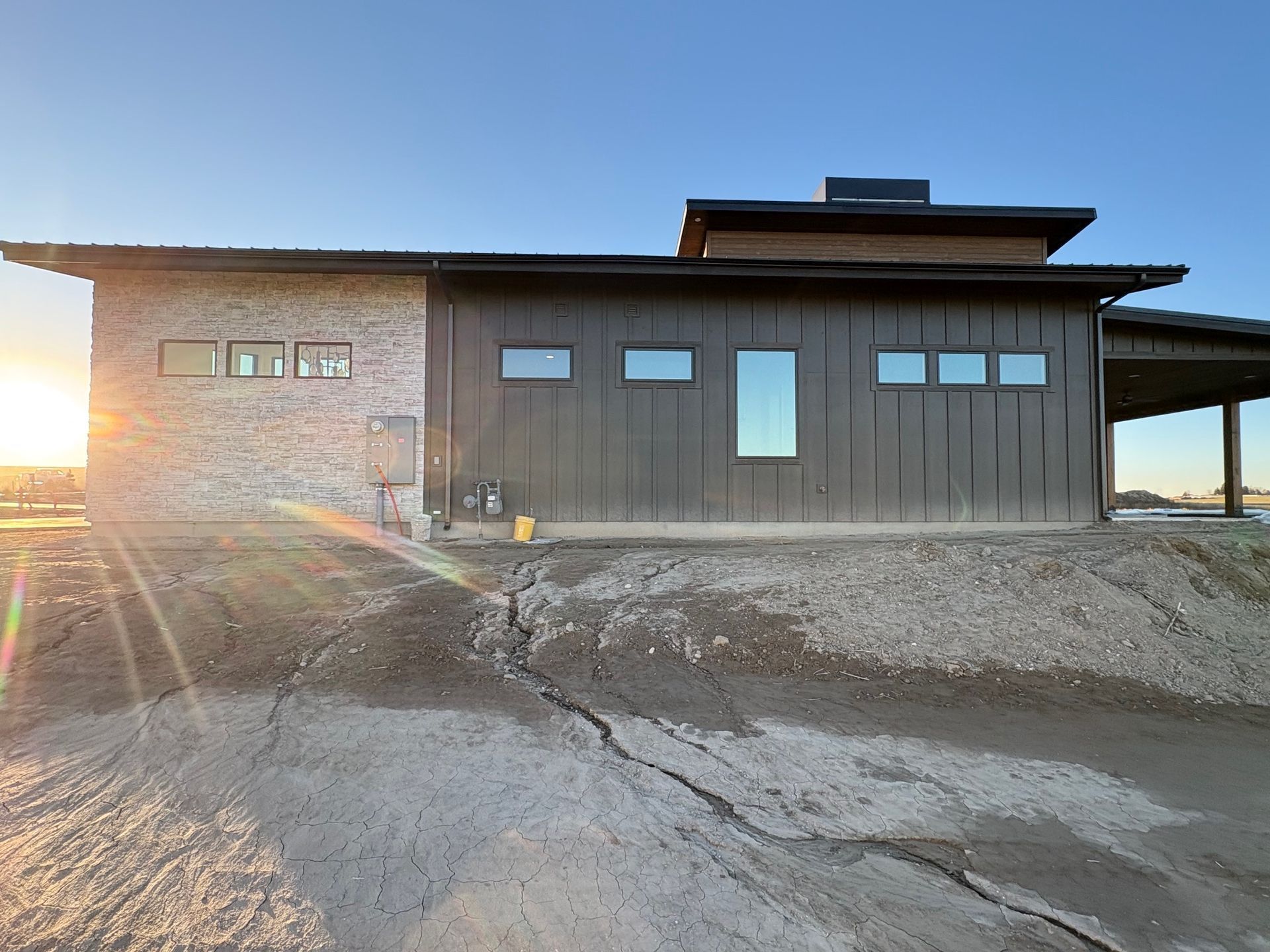Modern house exterior with brick and dark siding. Setting sun casts long shadows on the dirt.