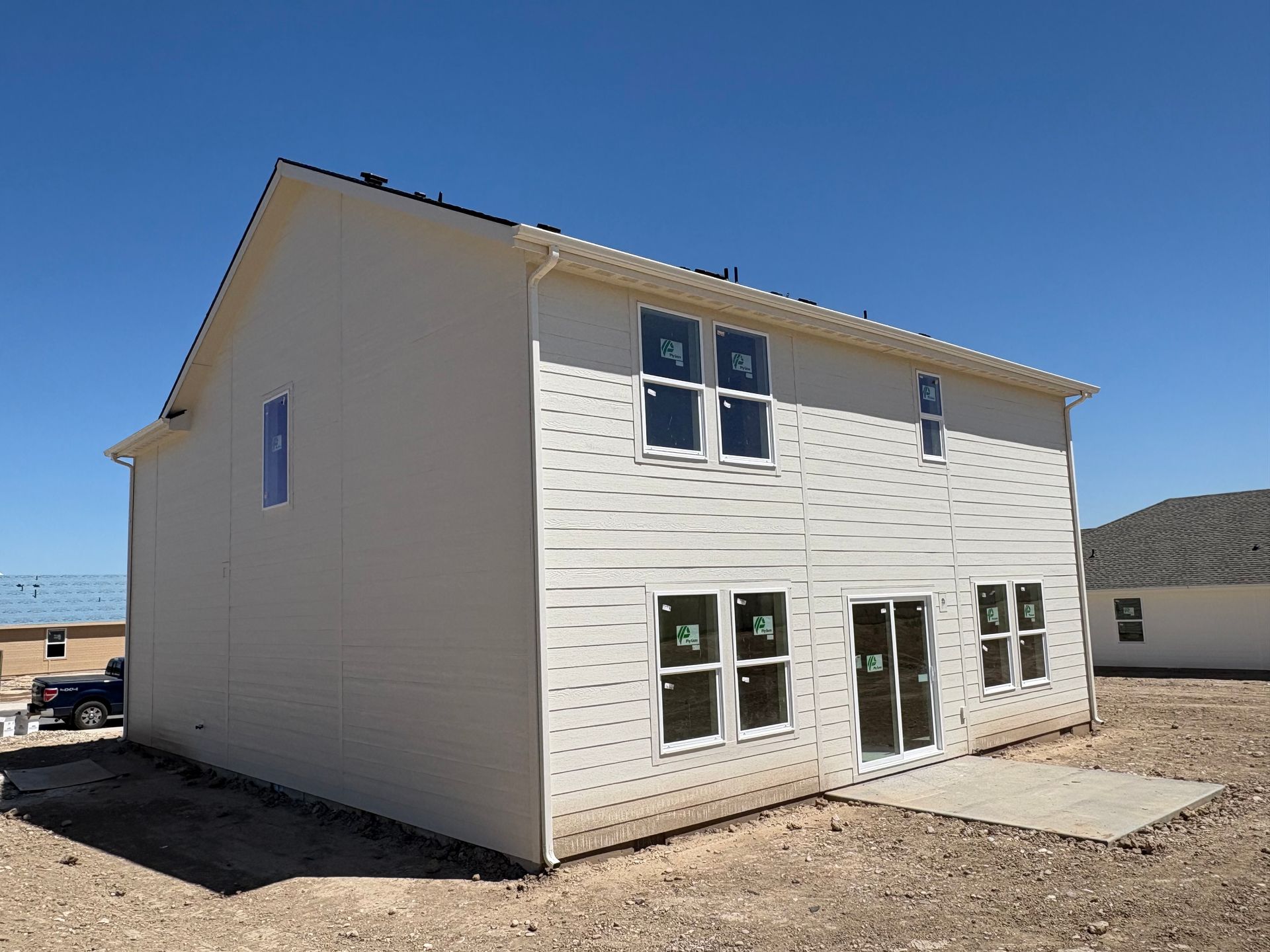 Back of a two-story beige house under construction with multiple windows and a concrete patio on a sunny day.