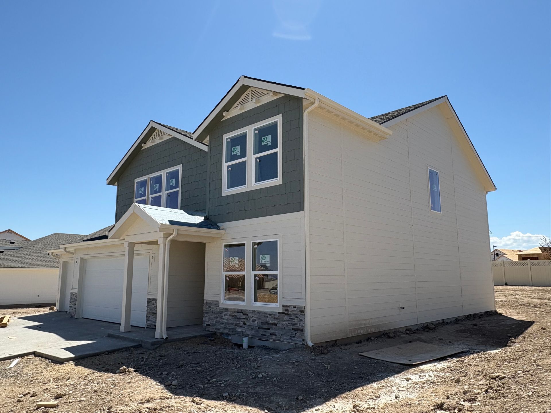 Two-story house under construction with light siding and blue-gray accent, surrounded by dirt under a blue sky.