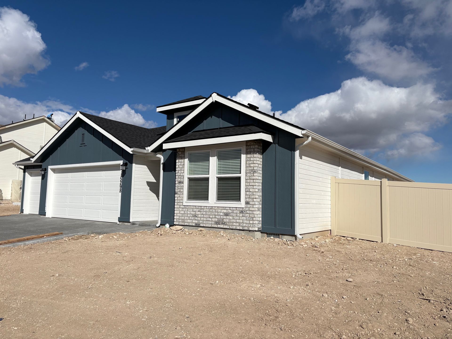 A modern blue and white house with a two-car garage under a blue sky, beige fencing, and dirt lot.