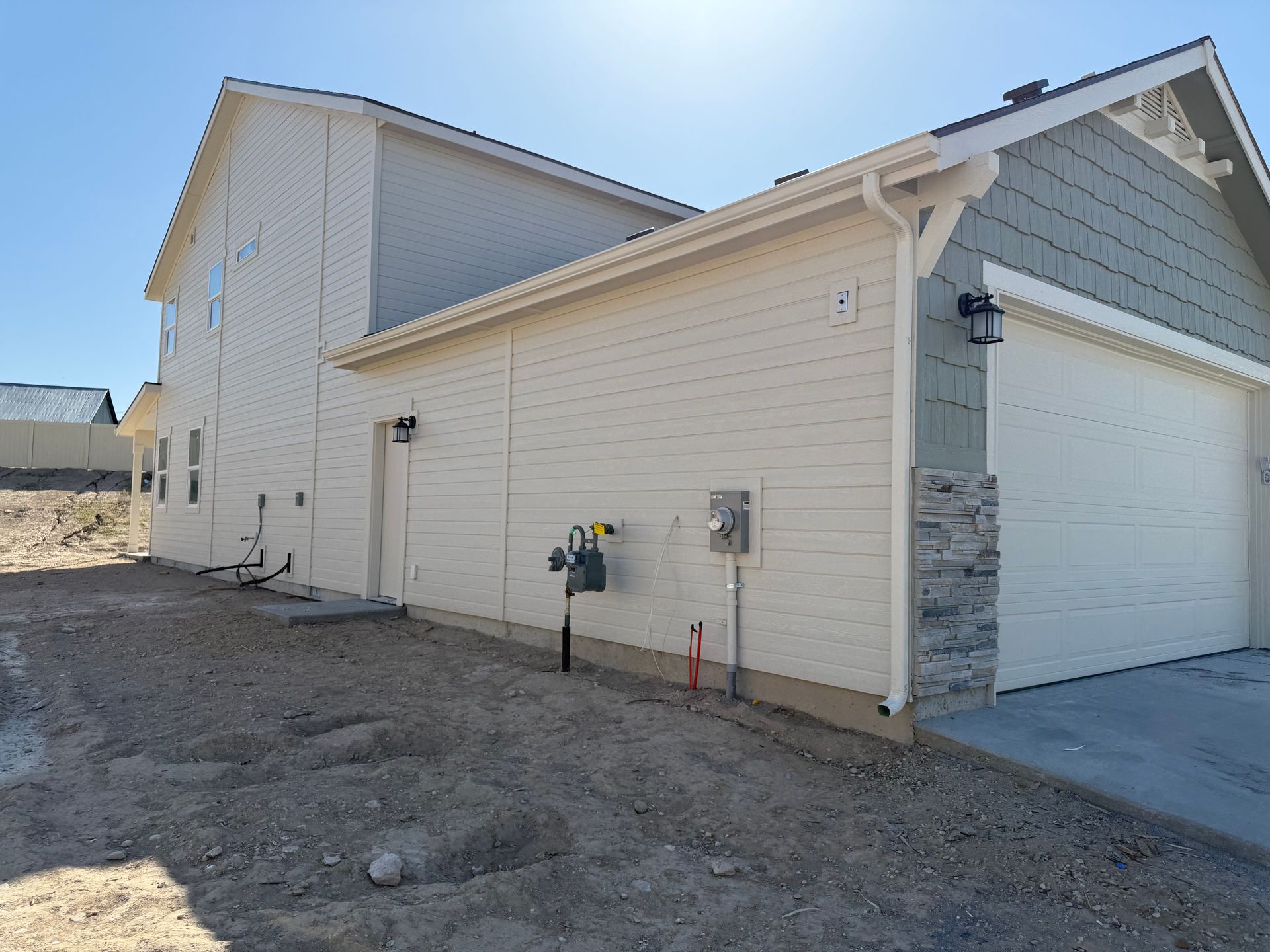 Side view of a two-story house with beige siding, garage, and gravel yard under a blue sky.
