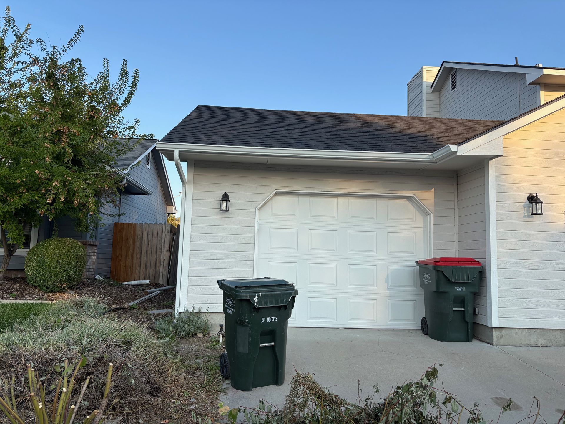 Garage with closed white door, green trash bins, shrubs, and a clear blue sky.