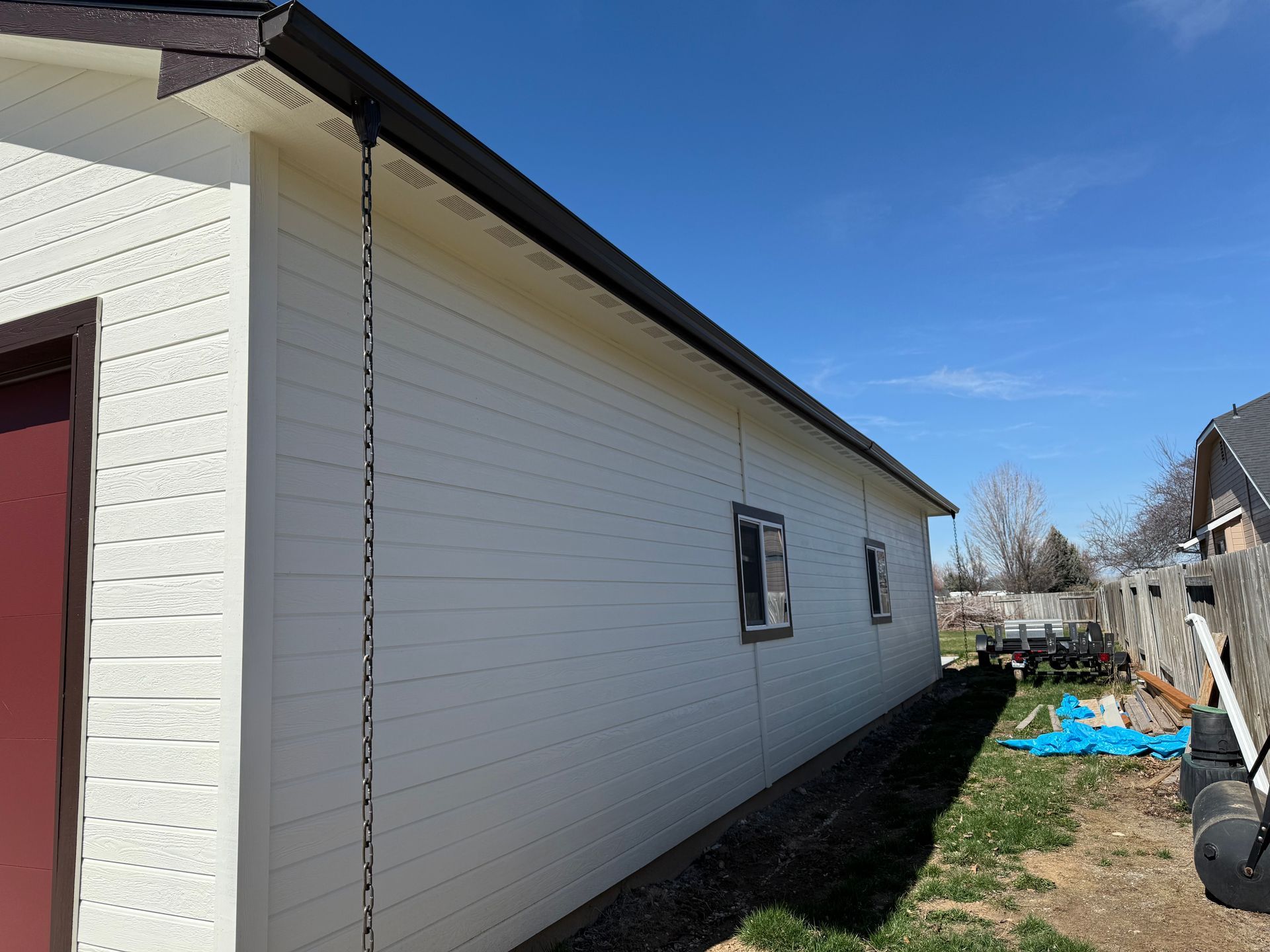 Off-white siding on a building with a dark brown roof and trim. A downspout chain hangs. Blue sky.