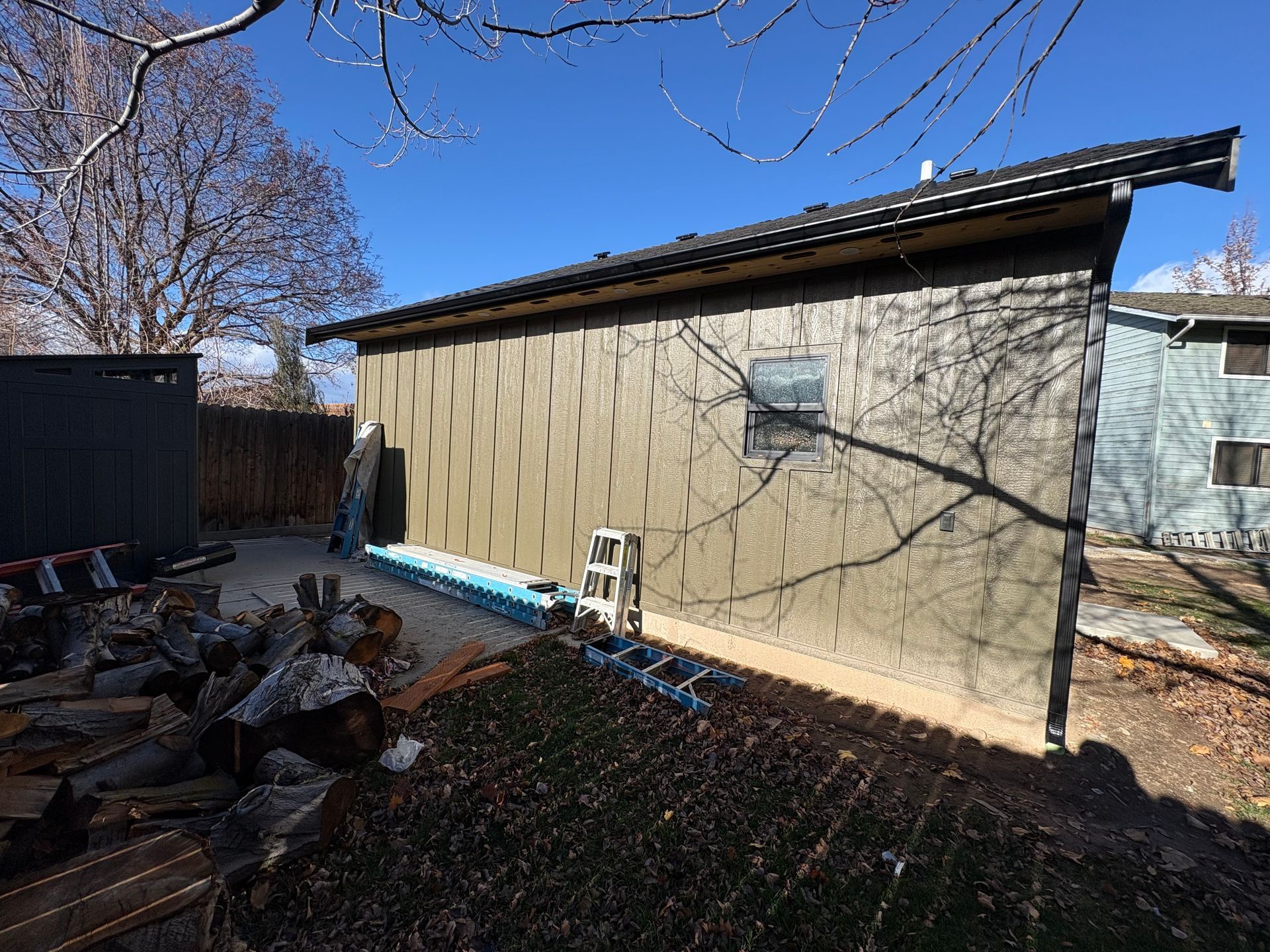Side view of a building with brown textured siding, small window, and a black roof. Construction materials present.