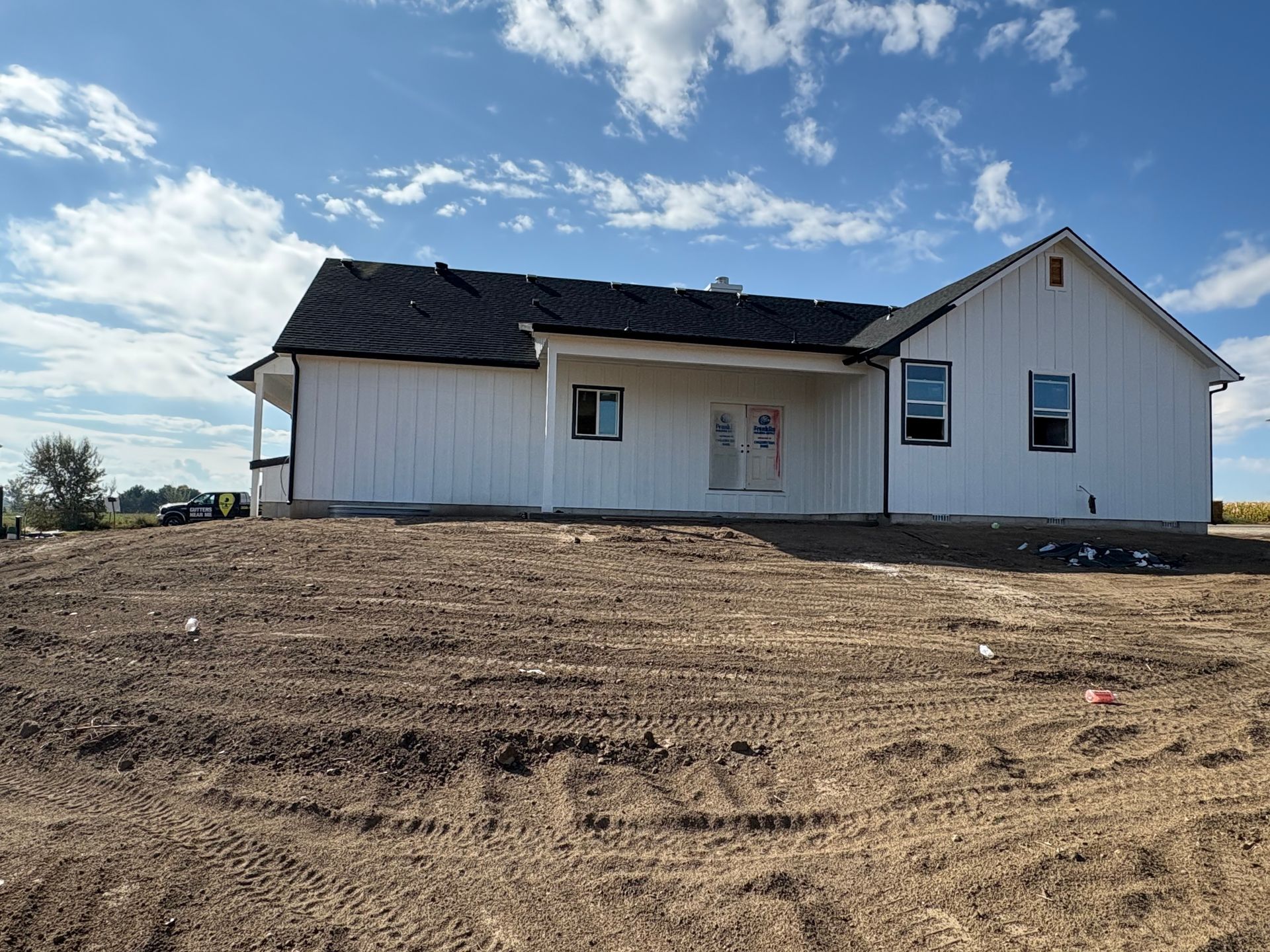 White house under construction with black roof, on a dirt lot, under blue sky with clouds.