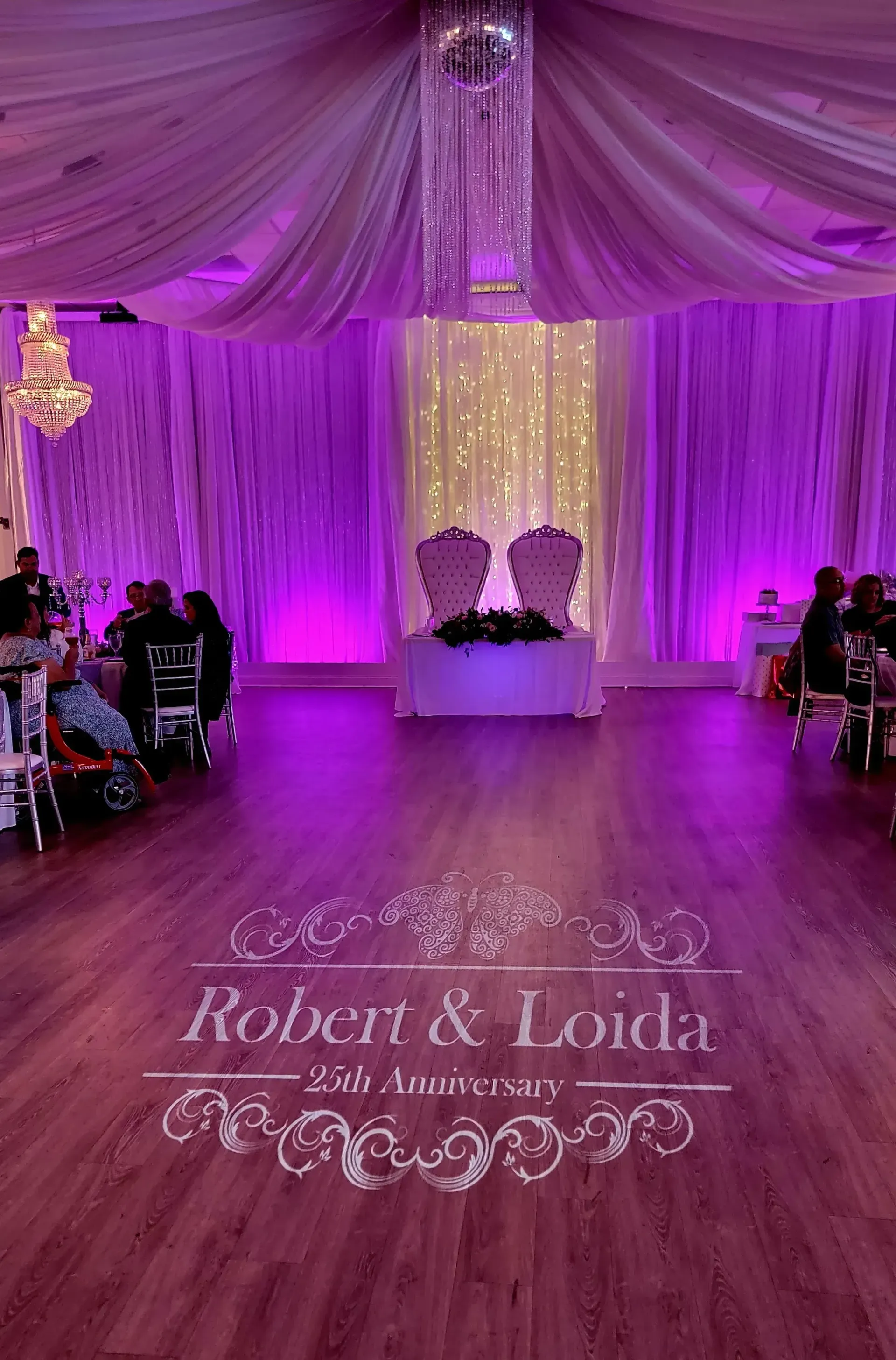 Purple-lit wedding reception hall with draped ceiling, sweetheart stage, and “Robert & Laila” dance floor monogram