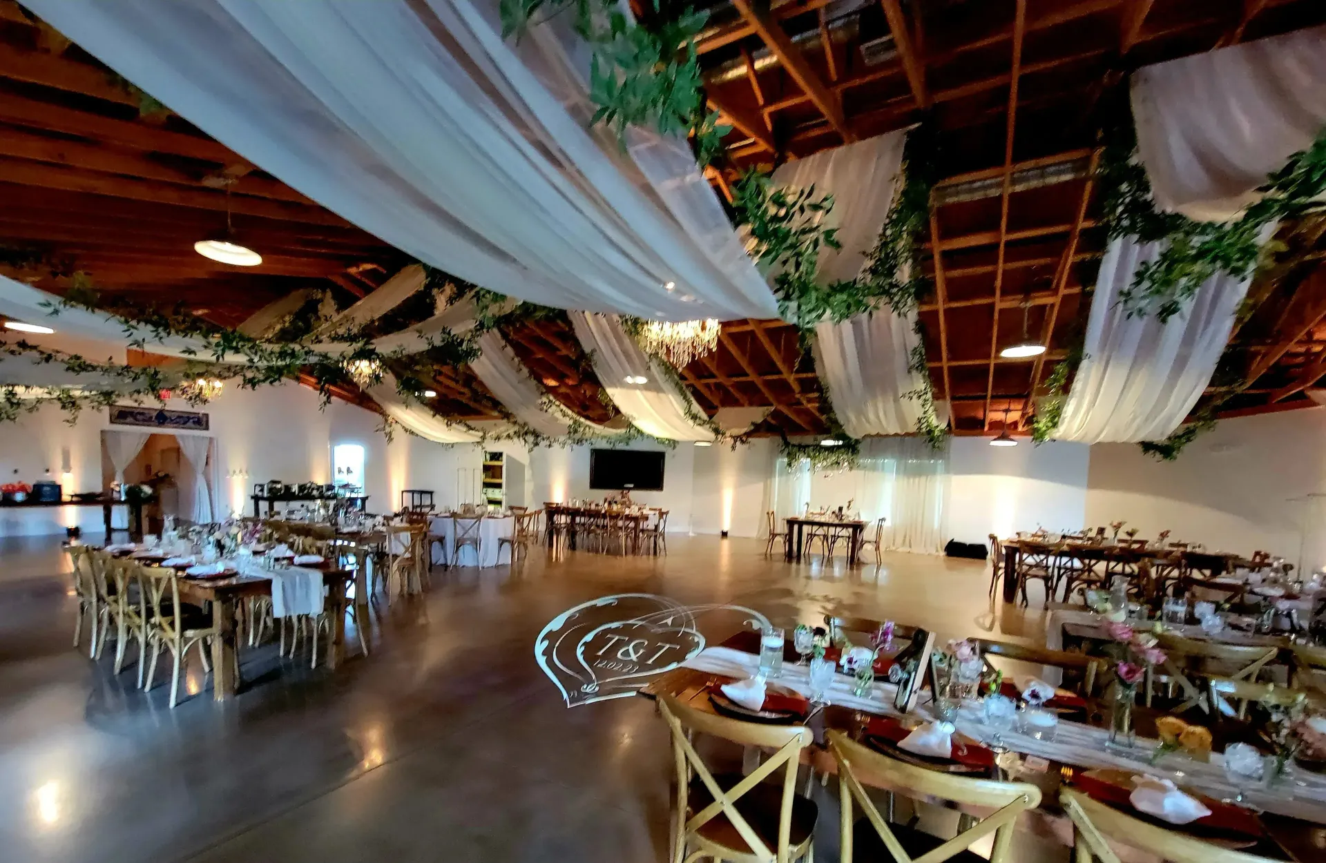 Rustic banquet hall with draped fabric, string lights, and set dining tables under a wooden roof