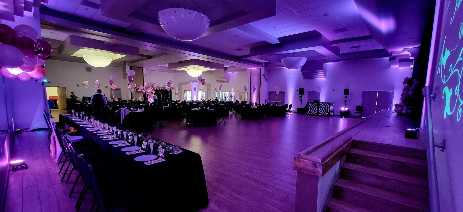 Purple-lit banquet hall with round tables, chairs, and a stage at the front