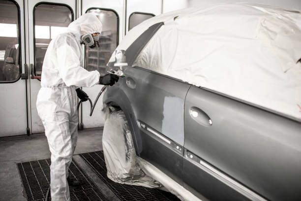 A man in a protective suit is painting a car in a garage.
