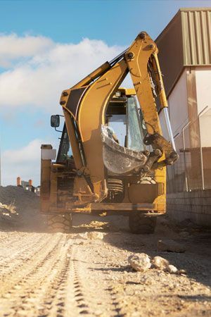 Yellow backhoe on a dirt road, scooping debris; building in the background.