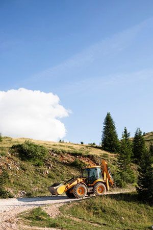 Yellow backhoe on a dirt road in a grassy hillside under a blue sky with a few clouds.