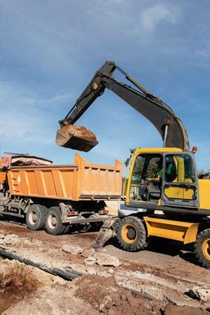 Yellow excavator loading an orange dump truck with dirt at a construction site under a blue sky.