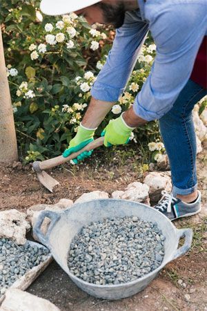 Person in garden using a hoe, with gravel in a bucket, wearing gloves and a hat.