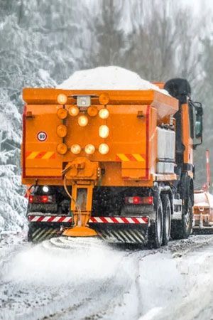 Orange snowplow truck spreading salt on a snow-covered road in a wintery forest.