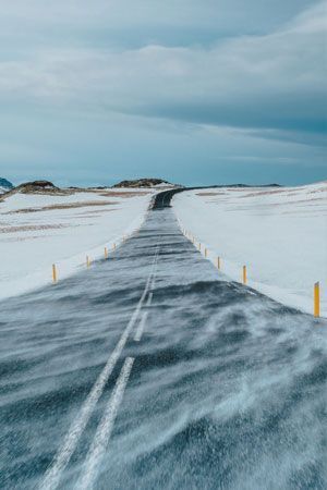 Road through snow-covered landscape with blowing snow, under a cloudy sky.