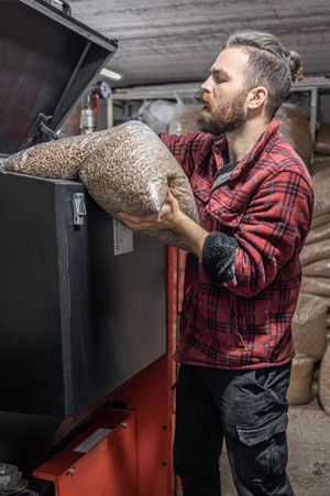 Man in red plaid shirt loading wood pellets into a heating unit; indoor setting.
