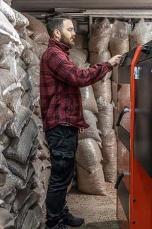 Man in red plaid shirt operating a pellet boiler, surrounded by bags of wood pellets in a storage room.