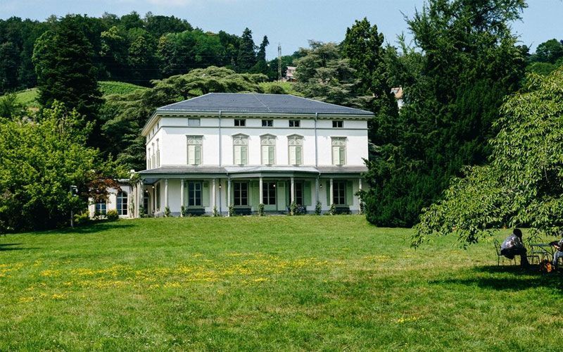 White, two-story house with dark roof in a green yard, trees in background, sunny day.