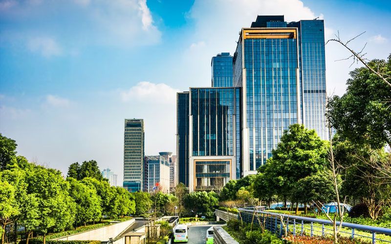 Modern city skyline with glass skyscrapers and lush green trees under a blue sky.