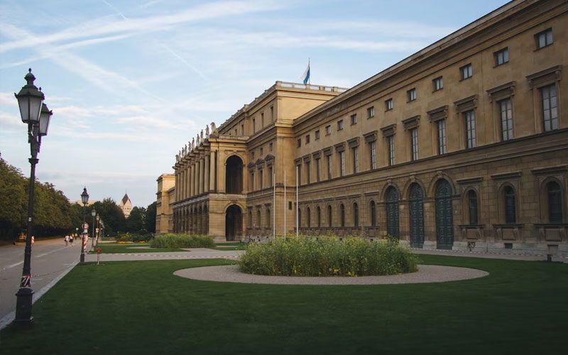 Long, light-colored building with arched entrances and many windows, green grass, and a lamp post; Munich, Germany.