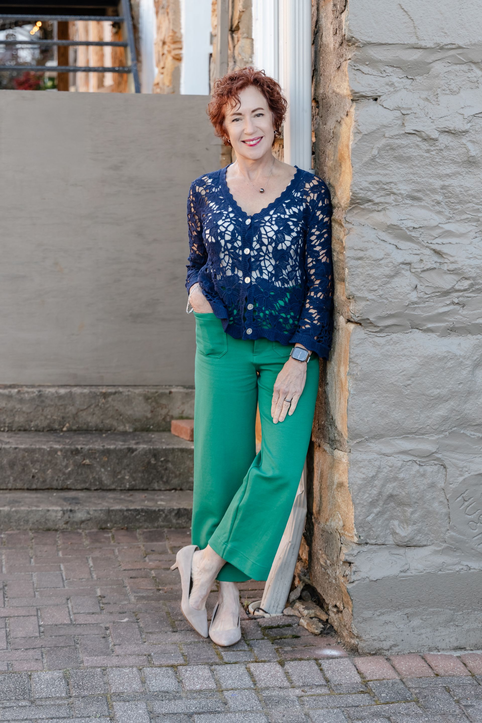 Woman in navy lace top and green pants leans against a stone wall, smiling.