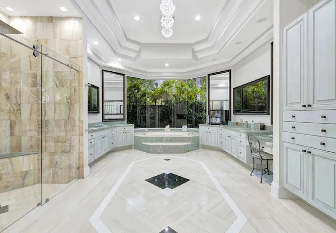 Spacious, light-filled bathroom with white cabinets, marble floors, a large window, and a glass shower.