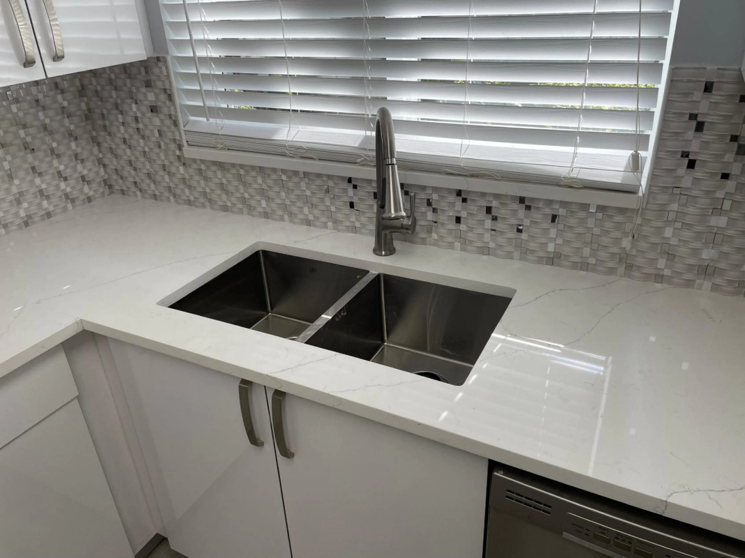 Bright white kitchen with double sink, stainless steel faucet, and tiled backsplash, under window with blinds.