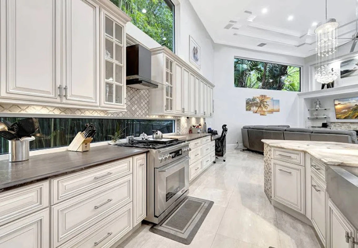 Elegant white kitchen with island, cabinetry, stainless steel appliances, and large windows overlooking lush greenery.