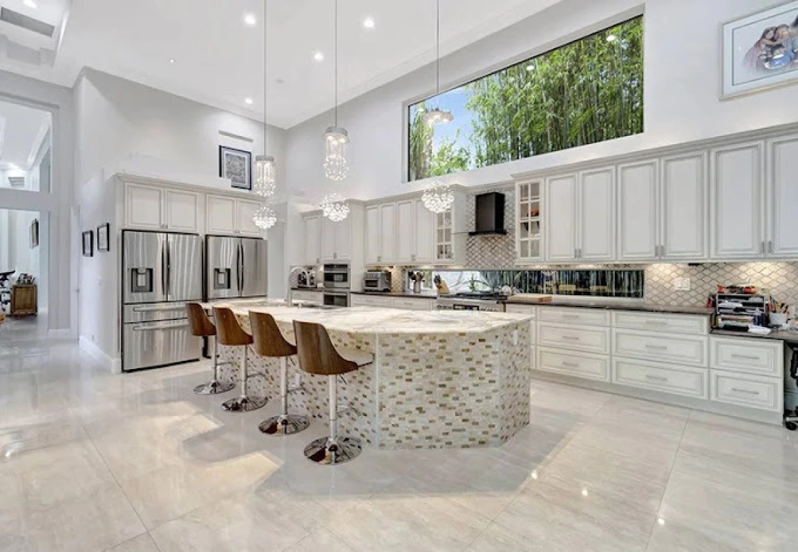 Spacious white kitchen with a large island, stainless steel appliances, and a tall window overlooking greenery.