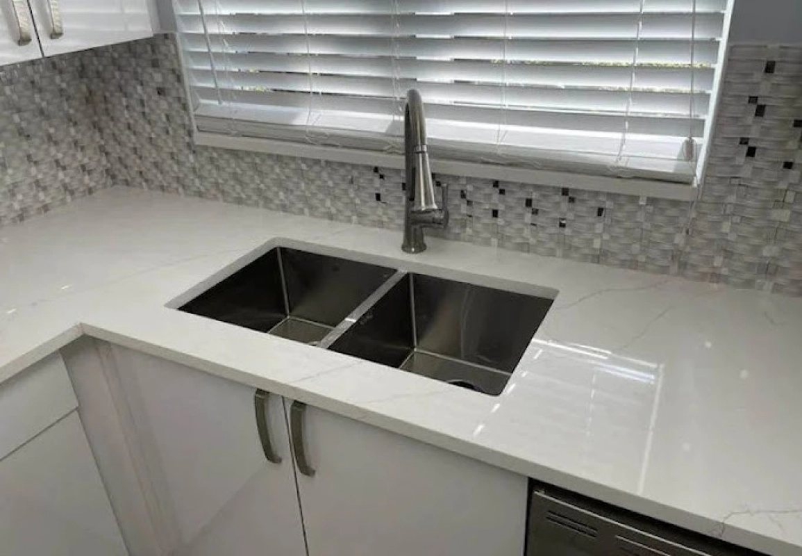 White kitchen countertop with stainless steel double sink and faucet. Blinds and mosaic tile backsplash visible.