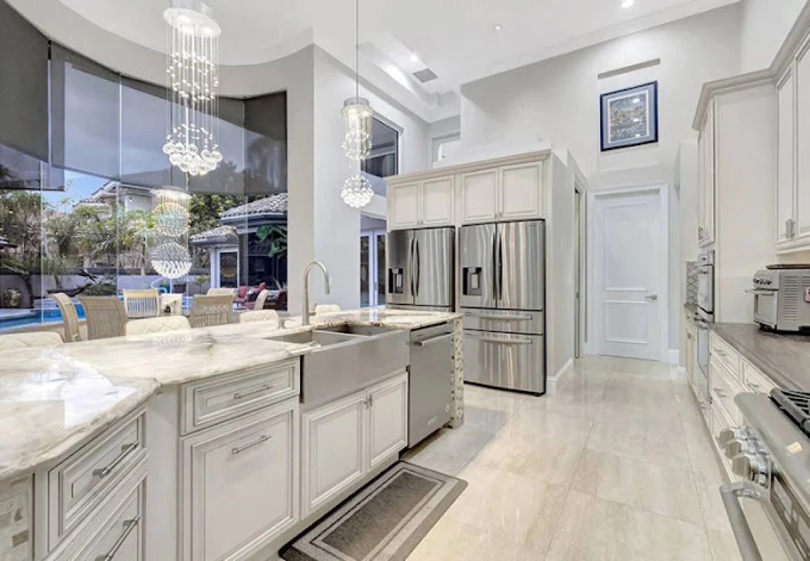Luxurious white kitchen with marble countertops, stainless steel appliances, and a large window overlooking a pool.