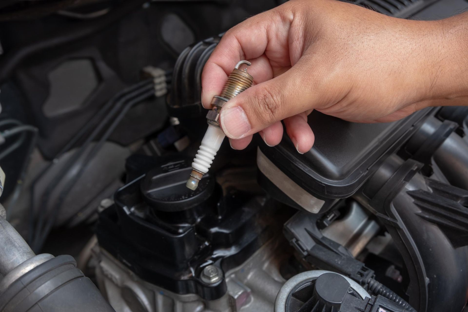 A person is holding a spark plug in their hand in front of a car engine.