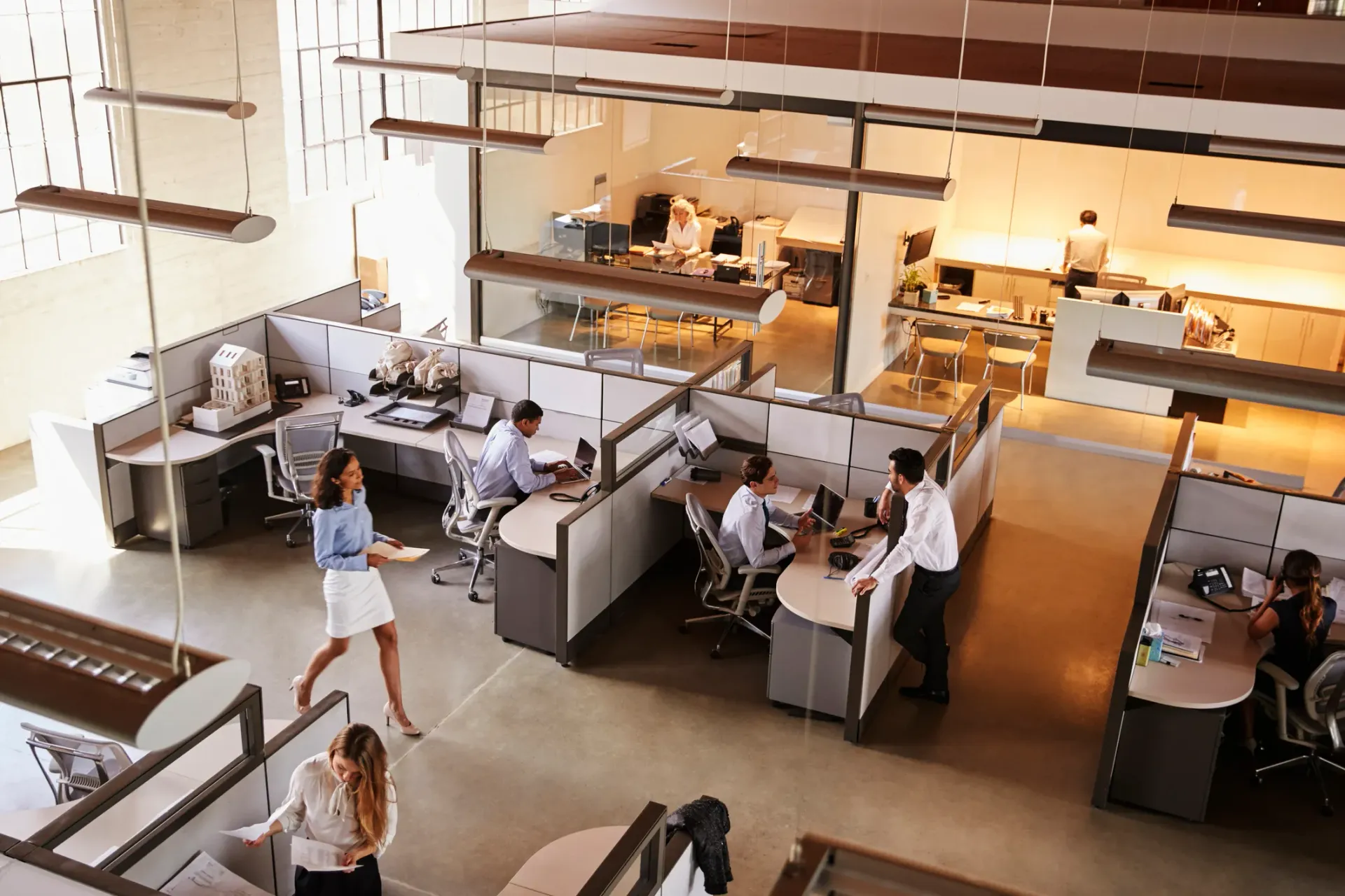 Busy office interior, workers at desks, woman walking, neutral tones, overhead view.