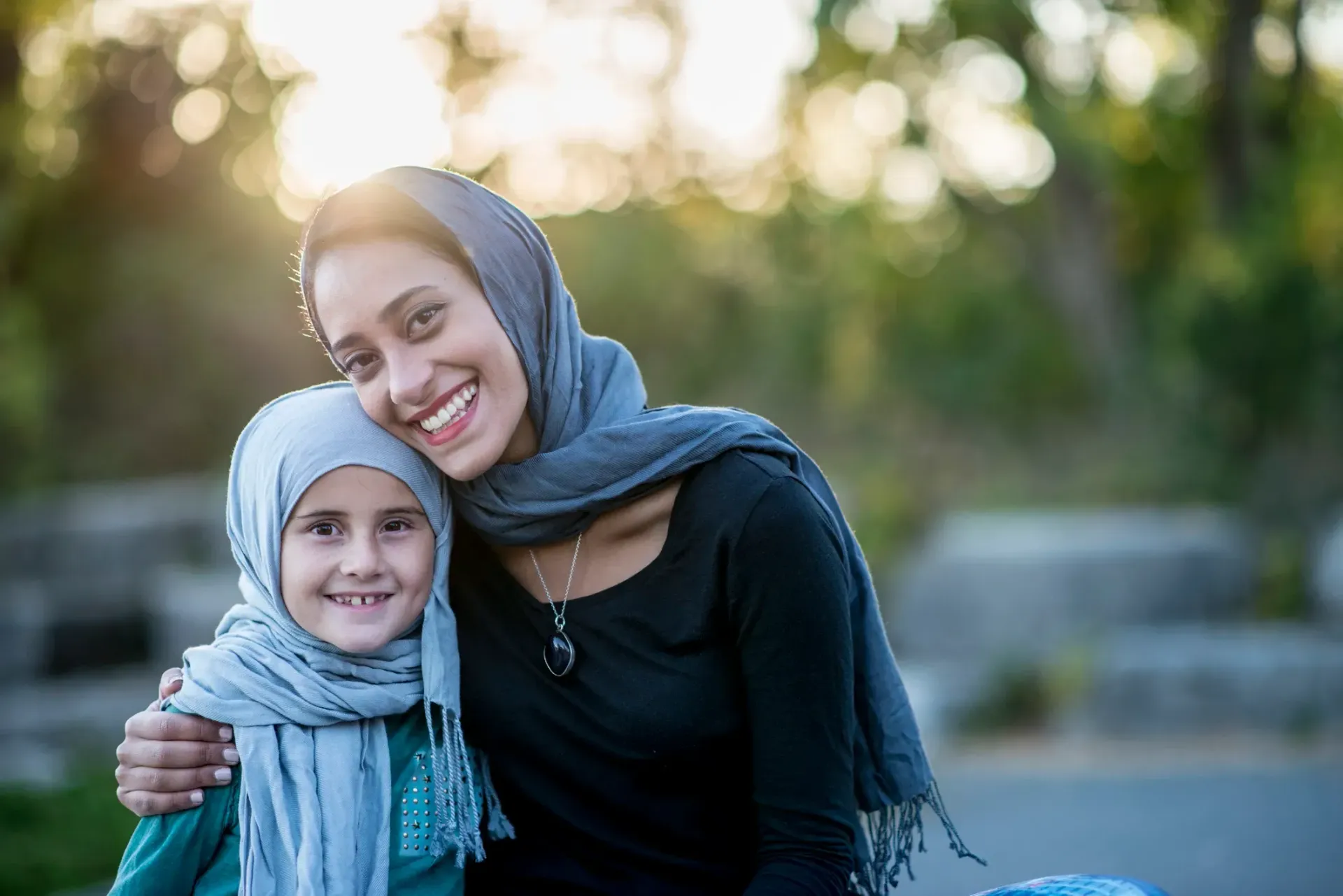 Woman in hijab smiling with a child in a hijab, outdoors in the sun.