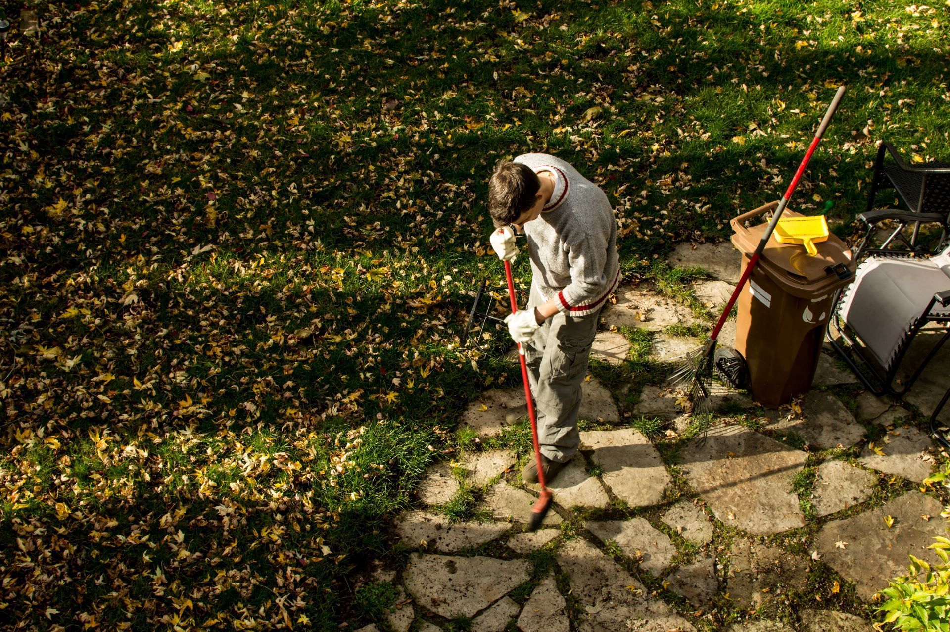 Man raking leaves on a patio with a trash can nearby.