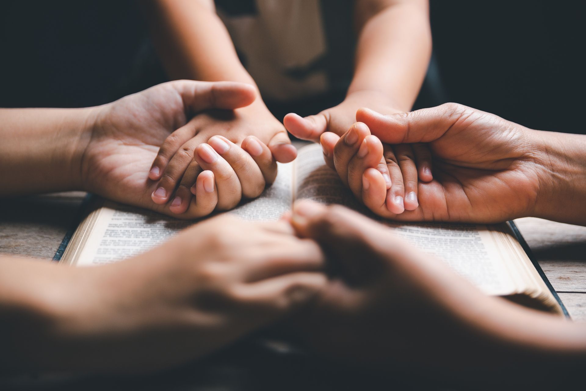 Hands of several people touching over an open book in a close, supportive circle.