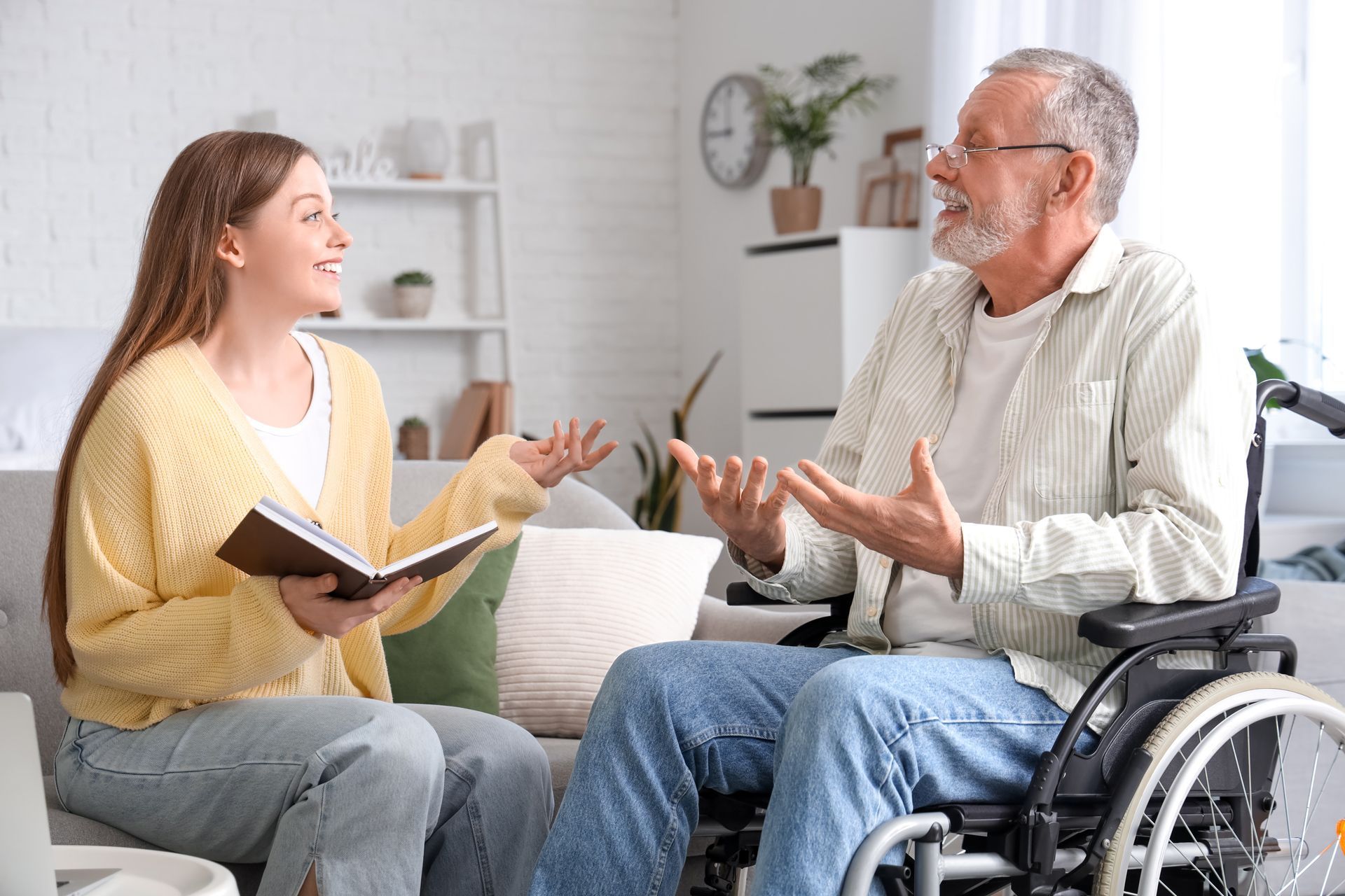 Two people chatting in a bright living room, one holding a notebook beside a man in a wheelchair