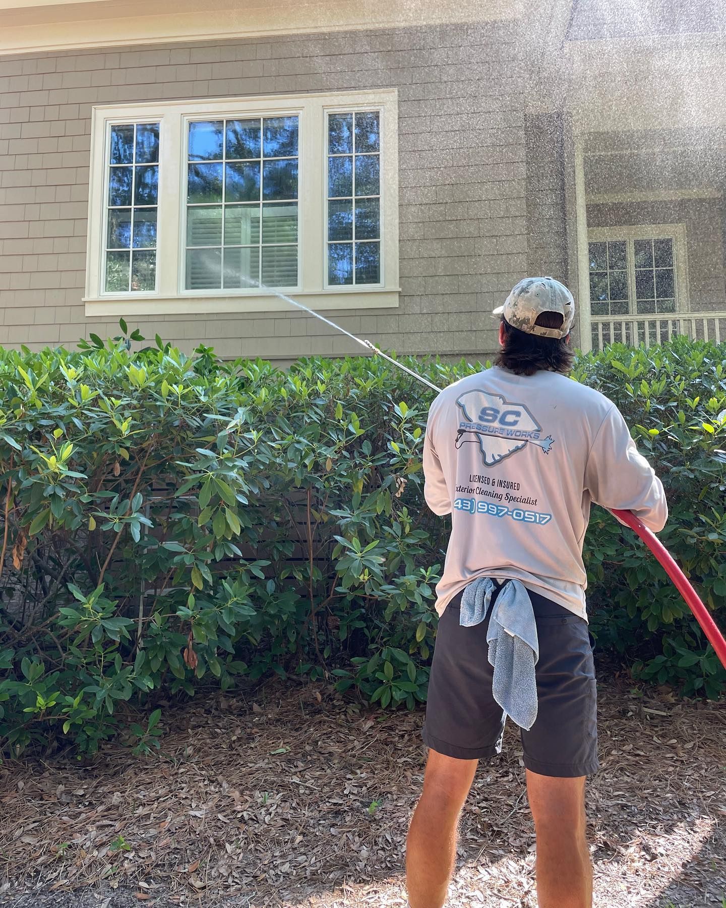Man pressure washing a house. Wearing a hat, tan shirt, and grey shorts. Outdoors, sunny day.