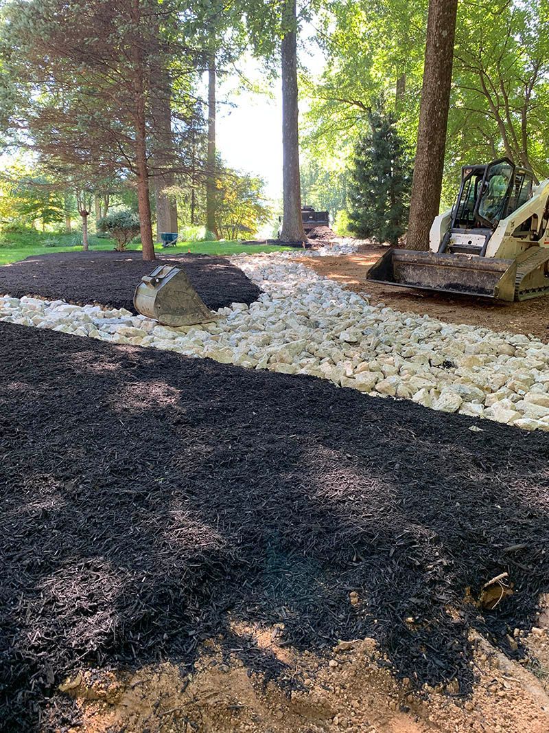 A pile of black mulch and rocks in a garden with trees in the background.