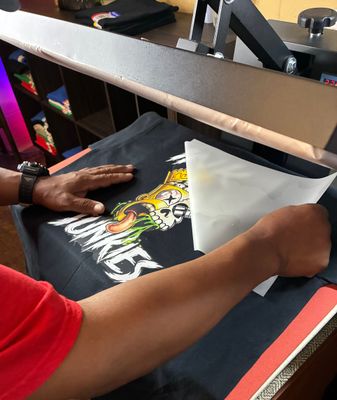 Hands arranging a white T-shirt with black printed text on a heat press table