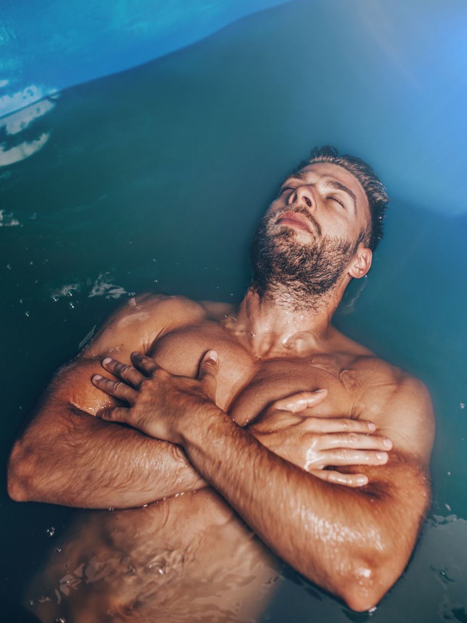 A shirtless man is laying in the water with his arms crossed.