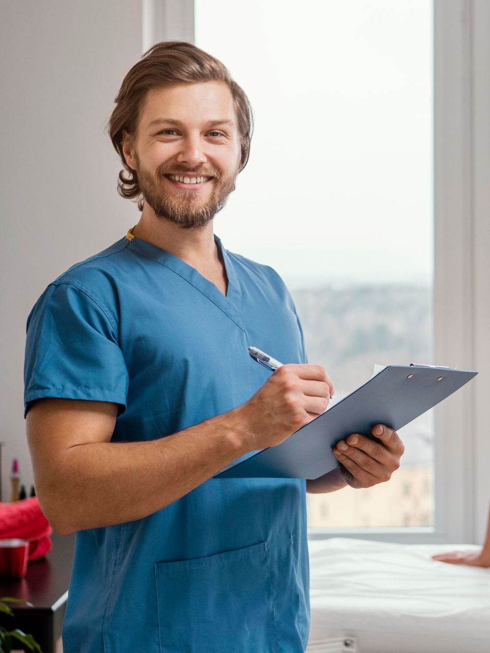 A man in a blue scrub is holding a clipboard and smiling.