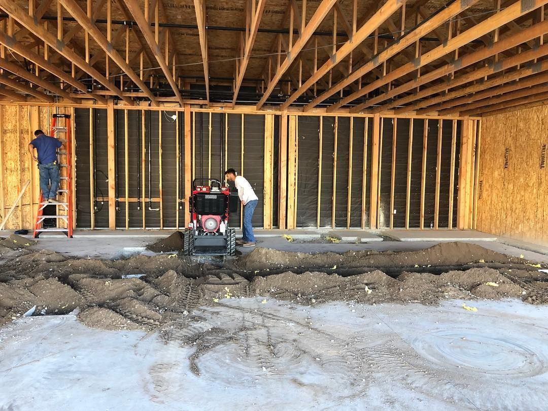 Construction site interior: two workers, one on a ladder, the other operating a machine, dirt mounds on the floor.