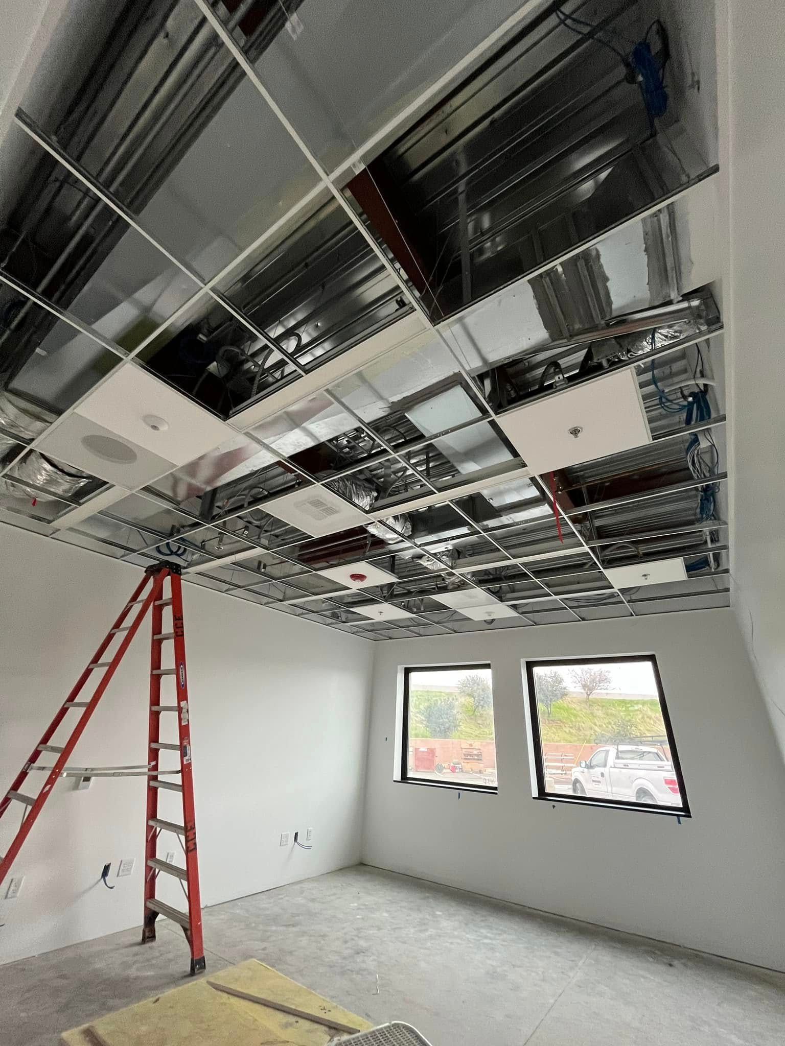 Interior room under construction; exposed metal ceiling grid, ladder, two windows.