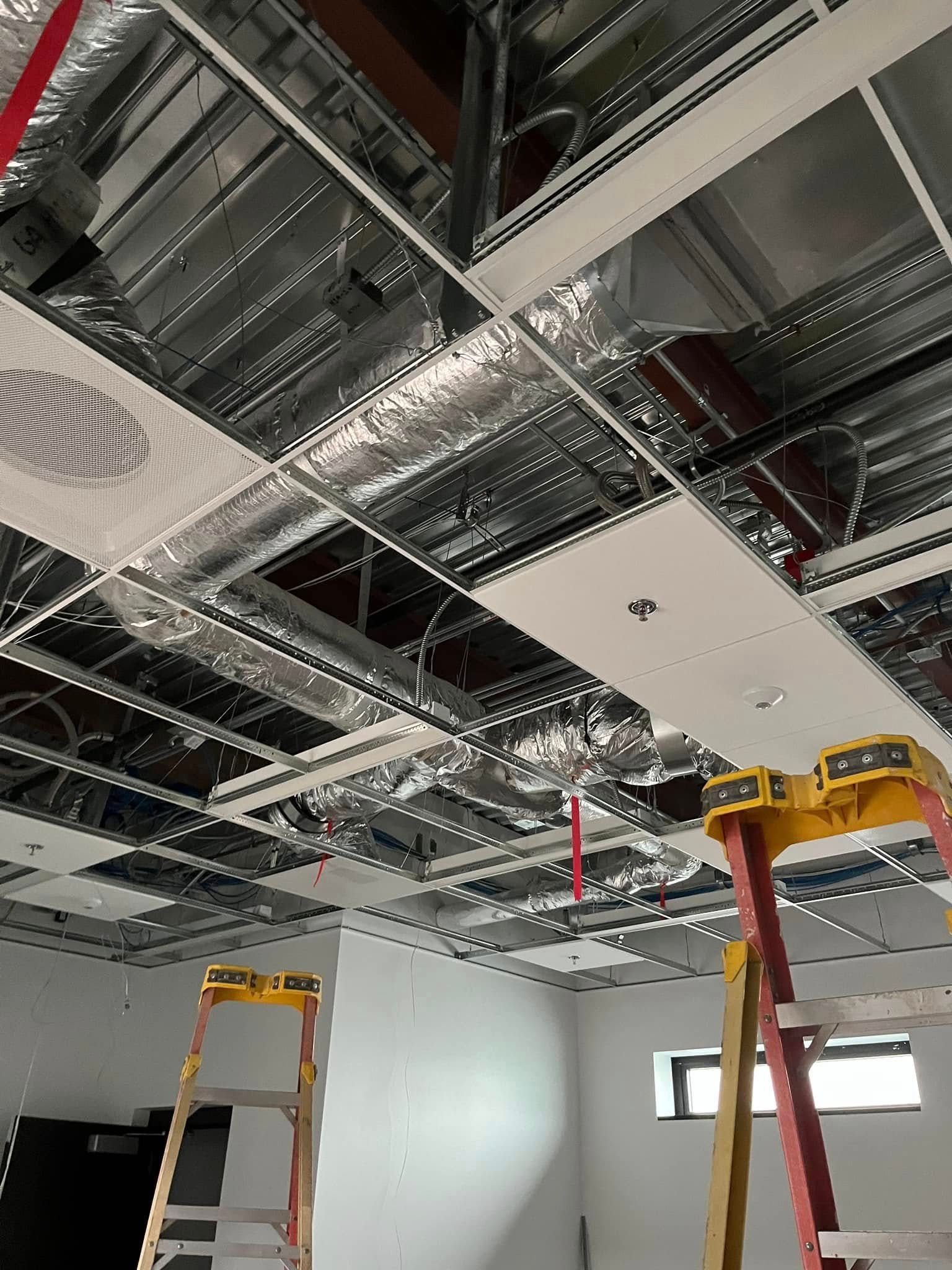 Construction of a commercial ceiling. Metal framework, ductwork, and partially installed panels. Two ladders are present.