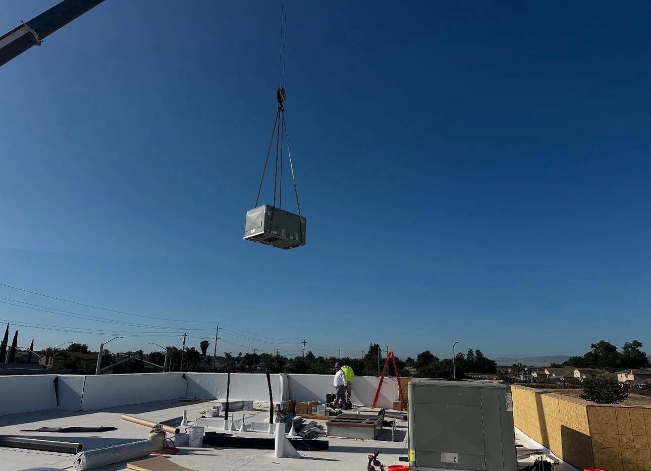 Crane lifting HVAC unit onto a white flat roof on a sunny day.
