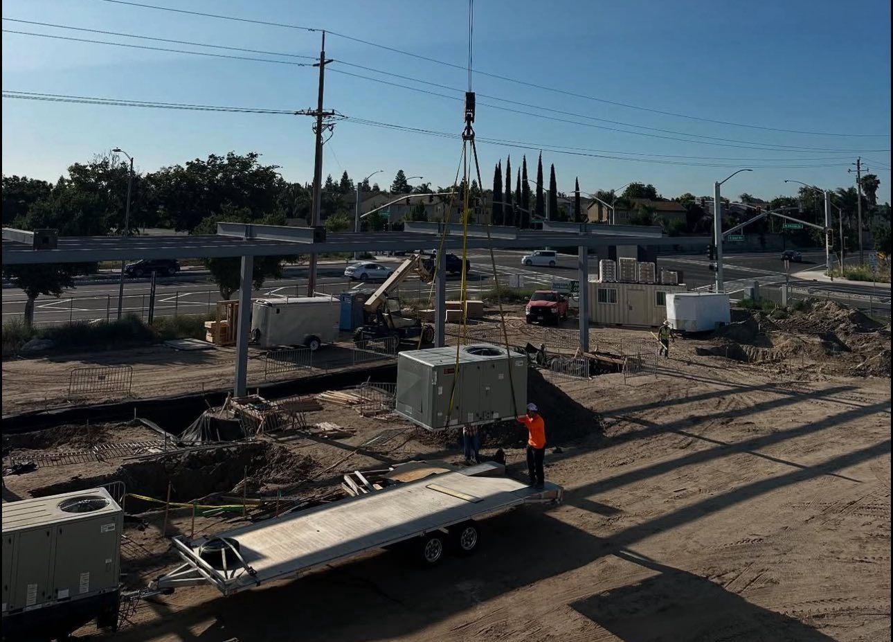 Construction site with a crane lifting a metal beam; worker near trailer.