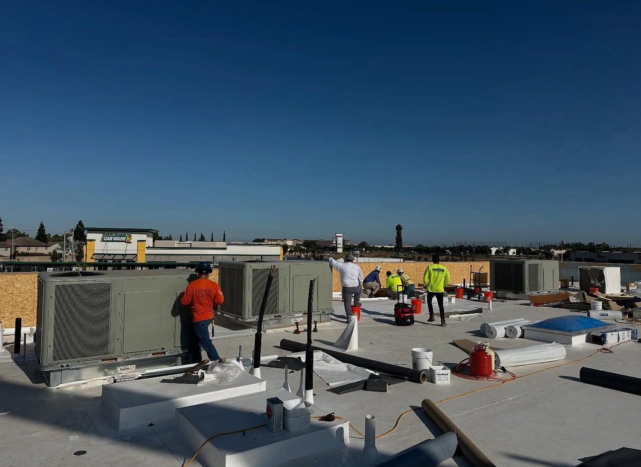 Construction workers on a flat rooftop with HVAC units, bright blue sky above.
