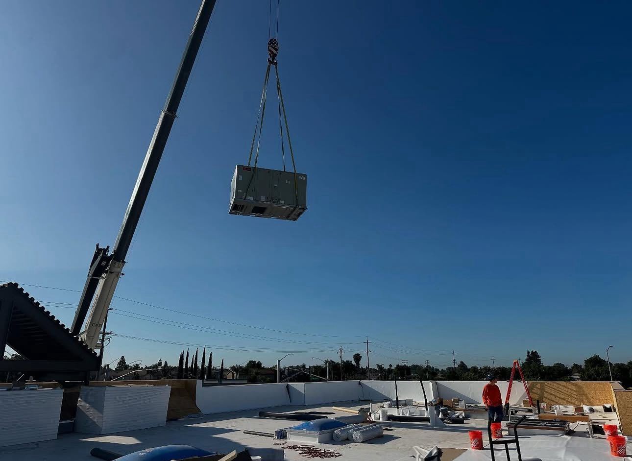 Crane lifting a large HVAC unit onto a flat roof. Construction workers are visible. Blue sky.