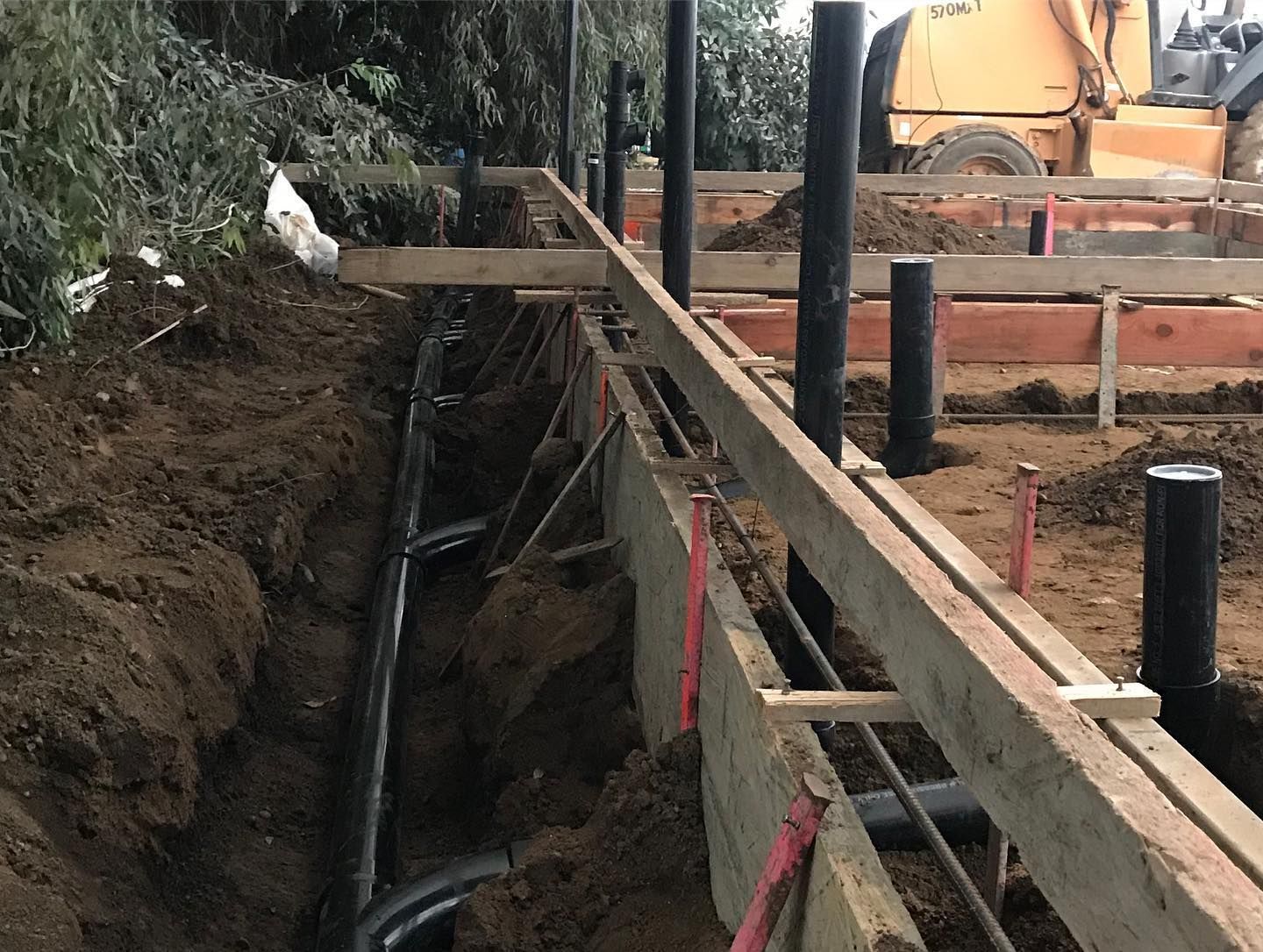 Construction site with wooden framework, exposed pipes in a trench, and earth-moving equipment in the background.